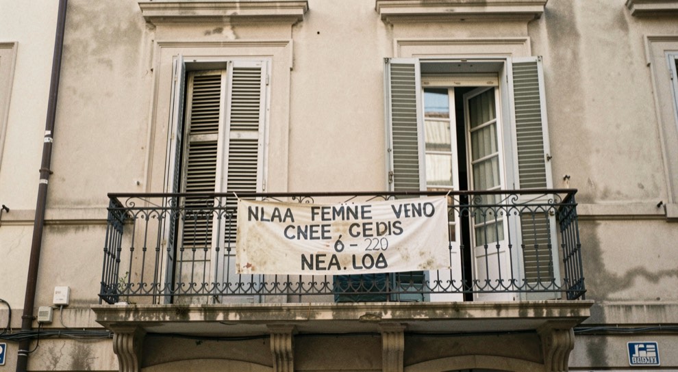 Facciata di una palazzina con due finestre chiuse dalle persiane e un cartello di vendita scolorito appeso al balcone