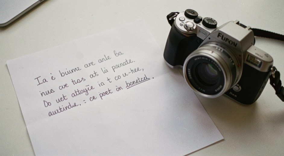 Foglio bianco con un testo di annuncio scritto a penna e corretto in due punti, accanto a una macchina fotografica compatta