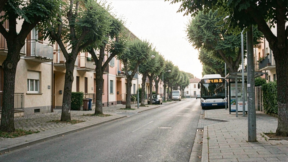 Strada di un quartiere residenziale italiano al mattino con palazzine basse, alberi lungo il marciapiede e una fermata dell'autobus