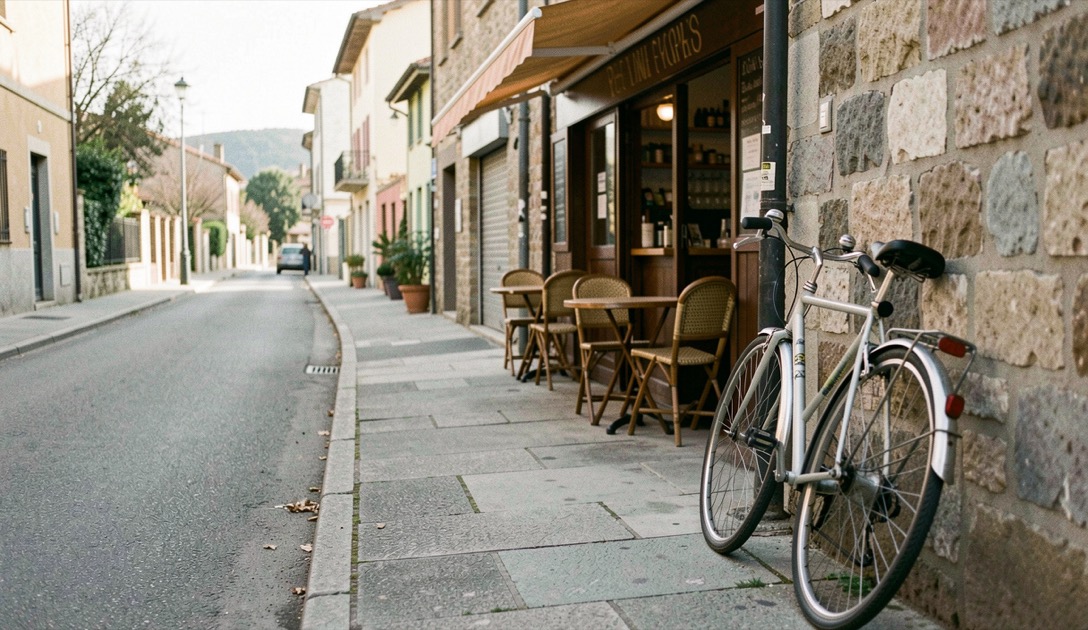 Marciapiede di una via di quartiere al mattino presto con un bar aperto, tavolini all'esterno e una bicicletta appoggiata al muro